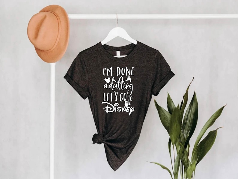 May include: Dark gray t-shirt with the text "I'M DONE adulting LET'S GO to Disney" in white script. The shirt is hanging on a white hanger. A brown hat hangs on a white pole to the left.