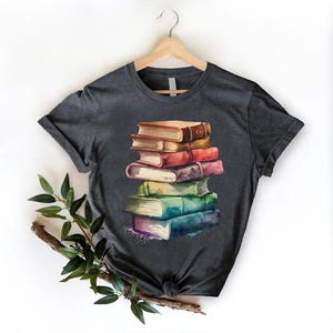 May include: Dark gray t-shirt featuring a watercolor illustration of a stack of books. The books are in various colors, including brown, red, purple, green, and blue. The shirt is hanging on a wooden hanger against a white background.