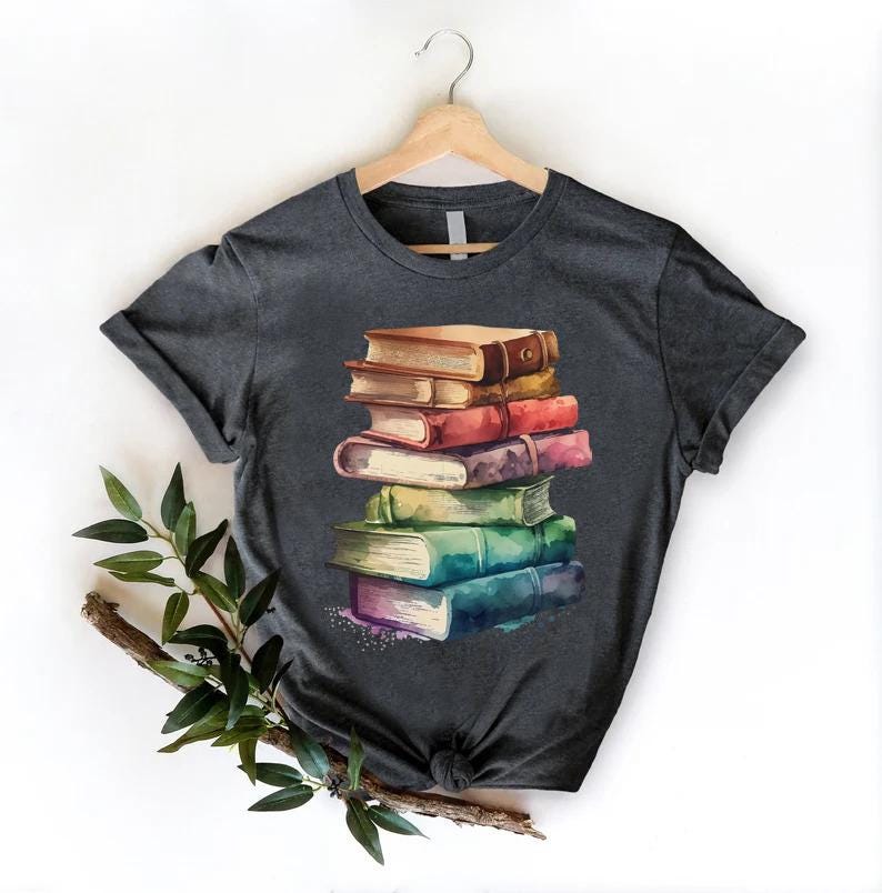 May include: Dark gray t-shirt featuring a watercolor illustration of a stack of books. The books are in various colors, including brown, red, purple, green, and blue. The shirt is hanging on a wooden hanger against a white background.