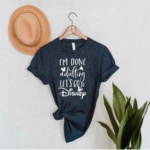 May include: Dark blue t-shirt with the text "I'M DONE adulting LET'S GO to Disney" in white. The shirt is hanging on a wooden hanger. A tan hat is to the left of the shirt.
