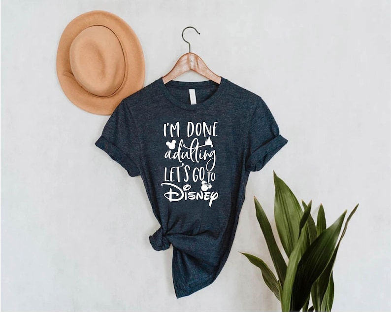 May include: Dark blue t-shirt with the text "I'M DONE adulting LET'S GO to Disney" in white. The shirt is hanging on a wooden hanger. A tan hat is to the left of the shirt.