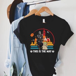 May include: Black t-shirt featuring a graphic of The Mandalorian, Grogu, and other Star Wars characters. The design includes a sunset background with the text "THIS IS THE WAY" in white. The shirt is hanging on a wooden hanger.