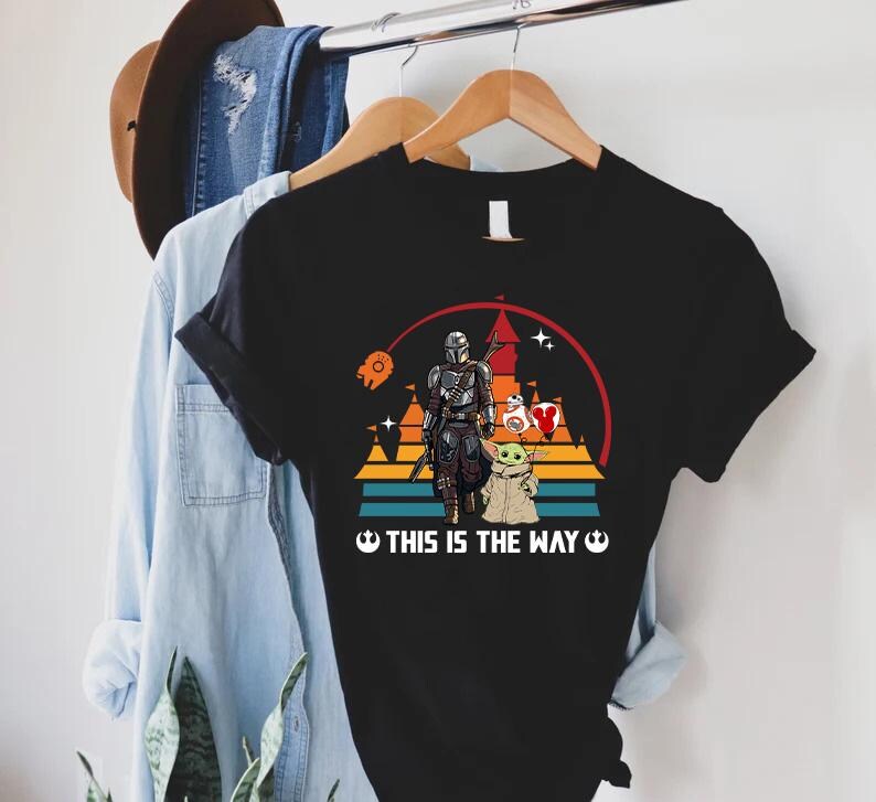 May include: Black t-shirt featuring a graphic of The Mandalorian, Grogu, and other Star Wars characters. The design includes a sunset background with the text "THIS IS THE WAY" in white. The shirt is hanging on a wooden hanger.