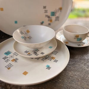 May include: A set of off-white ceramic dinnerware featuring a pattern of small black geometric line drawings, teal squares, and mustard yellow squares. The collection includes a large platter, a smaller plate, a bowl, and a teacup with a saucer.
