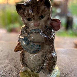 May include: A ceramic figurine of a cat holding a blue basket with a small brown duck. The cat has brown and gray fur with a pink nose and is standing on a green and yellow base. The figurine is detailed and has a vintage aesthetic.