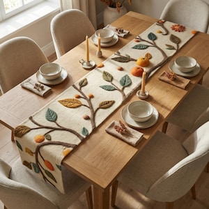 Op de afbeelding: Een houten eettafel gedekt voor een maaltijd, met een crèmekleurige tafelloper met een viltboomontwerp. De tafel is gedekt met kommen, borden, servetten, kaarsen en stoelen. De loper heeft groene, oranje en bruine bladeren en fruit.