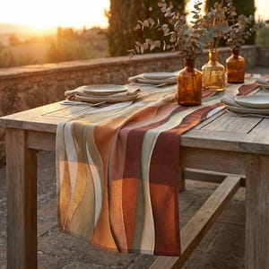 May include: A wooden table set for dining outdoors, featuring a colorful runner with wavy stripes in shades of beige, gold, and brown. The table is set with plates, silverware, and amber-colored vases with greenery.