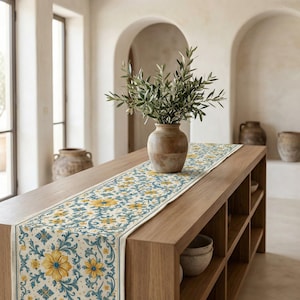 May include: A floral table runner with a cream background and yellow and teal flower pattern. The runner is placed on a wooden table, with a vase of greenery in the center. The table has built-in shelves.