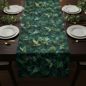 May include: A dark green table runner with a fern leaf pattern. The runner is placed on a dark brown wooden table set for a meal, with white plates, gold silverware, and clear wine glasses.