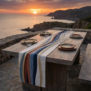 May include: A wooden table set for dining with a colorful runner. The runner features vertical stripes in shades of white, blue, gold, and brown. The table is set with plates and glasses. The background shows a sunset over the ocean.