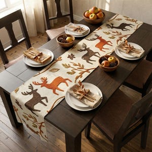Op de afbeelding: Een donkerhouten eettafel gedekt voor een maaltijd, met een crèmekleurige tafelloper met bruine en oranje hertenontwerpen. De tafel is gedekt met witte borden, servetten, houten keukengerei en kommen met appels en dennenappels.
