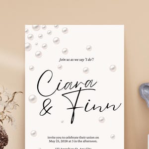 Può includere: Un invito di nozze bianco con un design a perle. L'invito recita "Unisciti a noi mentre diciamo 'Sì, lo voglio!' Ciara & Finn" e include la data, l'ora e il luogo del matrimonio. Il testo "Ricevimento a seguire" è in fondo all'invito.