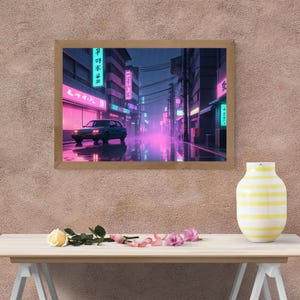 Puede incluir: Una impresión de arte digital enmarcada que representa una escena nocturna lluviosa en una ciudad japonesa. La calle está bordeada de edificios con letreros de neón en rosa y azul. Un coche blanco circula por la calle. La imagen tiene una estética retro, vaporwave.