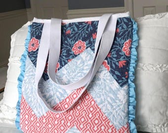 May include: A quilted tote bag featuring a chevron pattern with sections of dark blue floral print, light blue foliage print, and coral geometric print. The bag has white fabric handles and is trimmed with light blue ruffles on the sides.