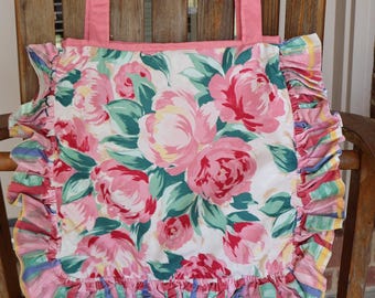 May include: A floral tote bag with pink handles and a ruffled edge. The bag features a white background with large pink roses, green leaves, and a colorful ruffled trim. The bag is sitting on a wooden chair.