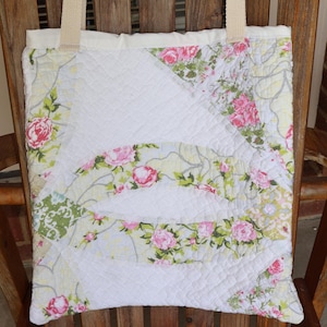 May include: A quilted tote bag with a floral pattern featuring pink roses and green leaves on a white background. The bag has cream-colored handles and is displayed on a wooden chair.