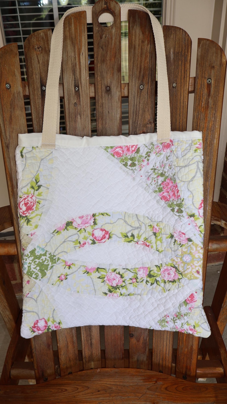May include: A quilted tote bag with a floral pattern featuring pink roses and green leaves on a white background. The bag has cream-colored handles and is displayed on a wooden chair.