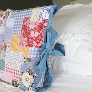 May include: A patchwork pillow with a variety of floral and gingham patterns in red, blue, yellow, and white. The pillow is trimmed with a light blue fabric and tied with a bow. The pillow is resting on a white bed.