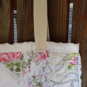 May include: Close-up of a quilted tote bag with a floral pattern. The bag features a cream-colored strap and a white border. The floral design includes pink roses and green leaves on a white background. The bag is hanging against a wooden fence.