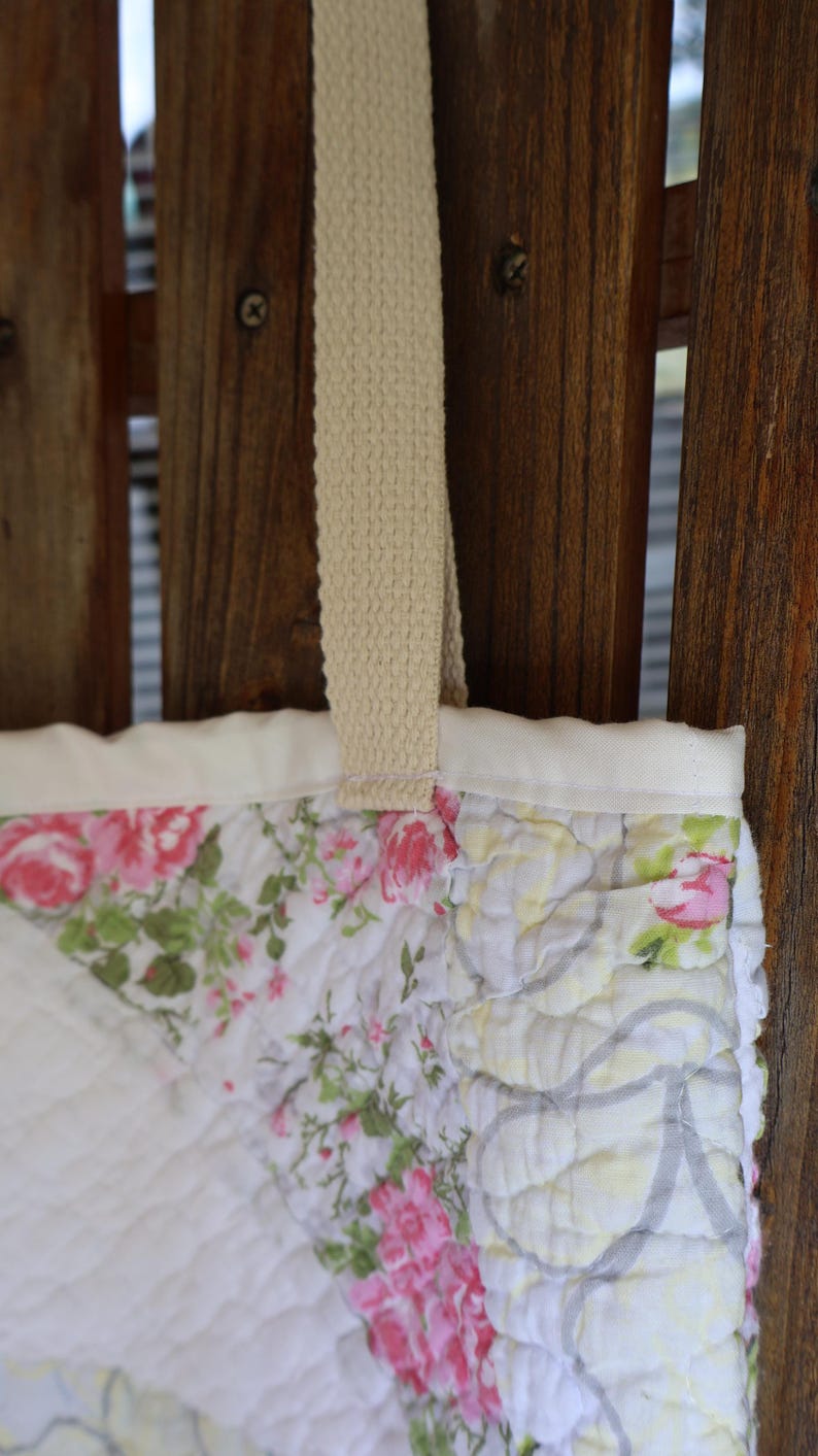 May include: Close-up of a quilted tote bag with a floral pattern. The bag features a cream-colored strap and a white border. The floral design includes pink roses and green leaves on a white background. The bag is hanging against a wooden fence.