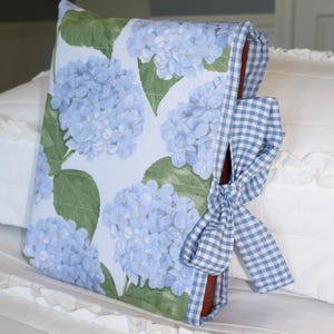 May include: A fabric book cover featuring a light blue hydrangea floral pattern with green leaves. The cover includes a blue and white gingham check fabric trim along the spine and edges, secured with a matching gingham ribbon tied in a bow.