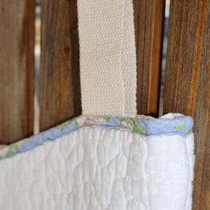 May include: Close-up of a white quilted tote bag with a floral patterned trim in pink, green, and blue. The bag has a cream-colored strap. The bag is hanging against a wooden background.