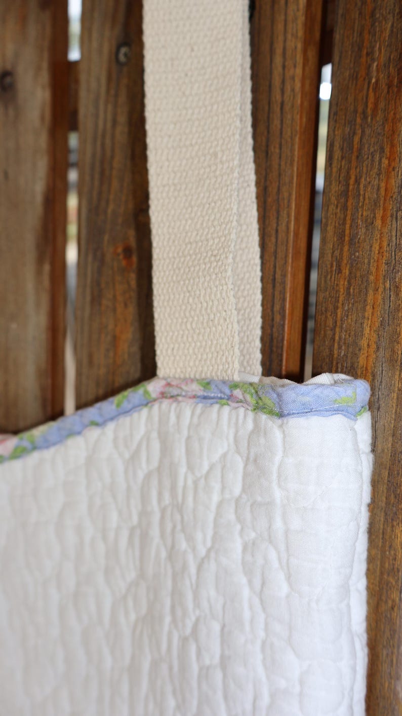 May include: Close-up of a white quilted tote bag with a floral patterned trim in pink, green, and blue. The bag has a cream-colored strap. The bag is hanging against a wooden background.