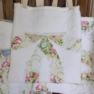 May include: White quilted tote bag with floral pocket detail. The bag features a quilted pattern and sturdy beige straps. The pocket has a floral pattern with pink roses and green leaves. The bag is sitting on a wooden chair.