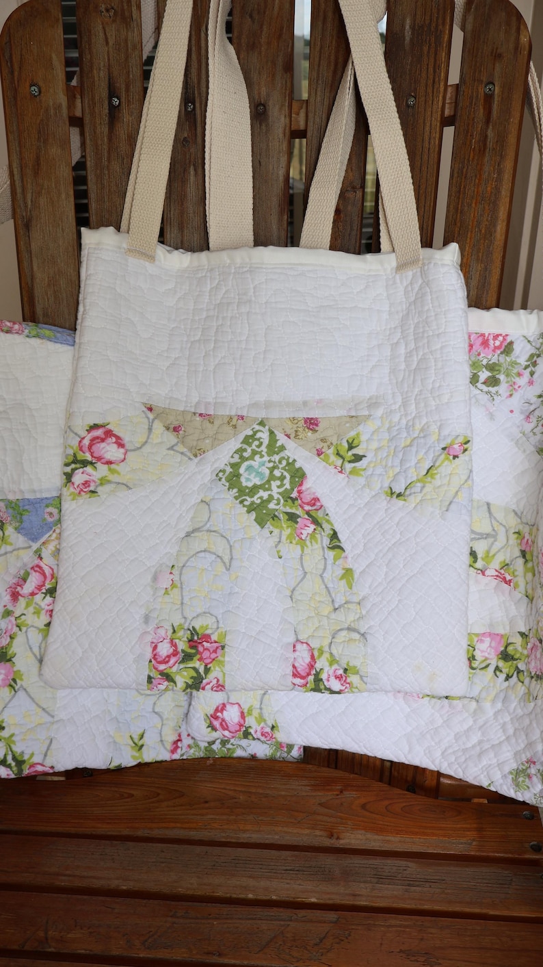 May include: White quilted tote bag with floral pocket detail. The bag features a quilted pattern and sturdy beige straps. The pocket has a floral pattern with pink roses and green leaves. The bag is sitting on a wooden chair.