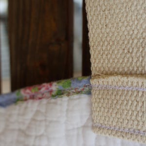 May include: Close-up of a quilted white fabric with a floral patterned edge and a beige woven strap. The strap is stitched to the fabric with a light purple thread. The background features a wooden structure.