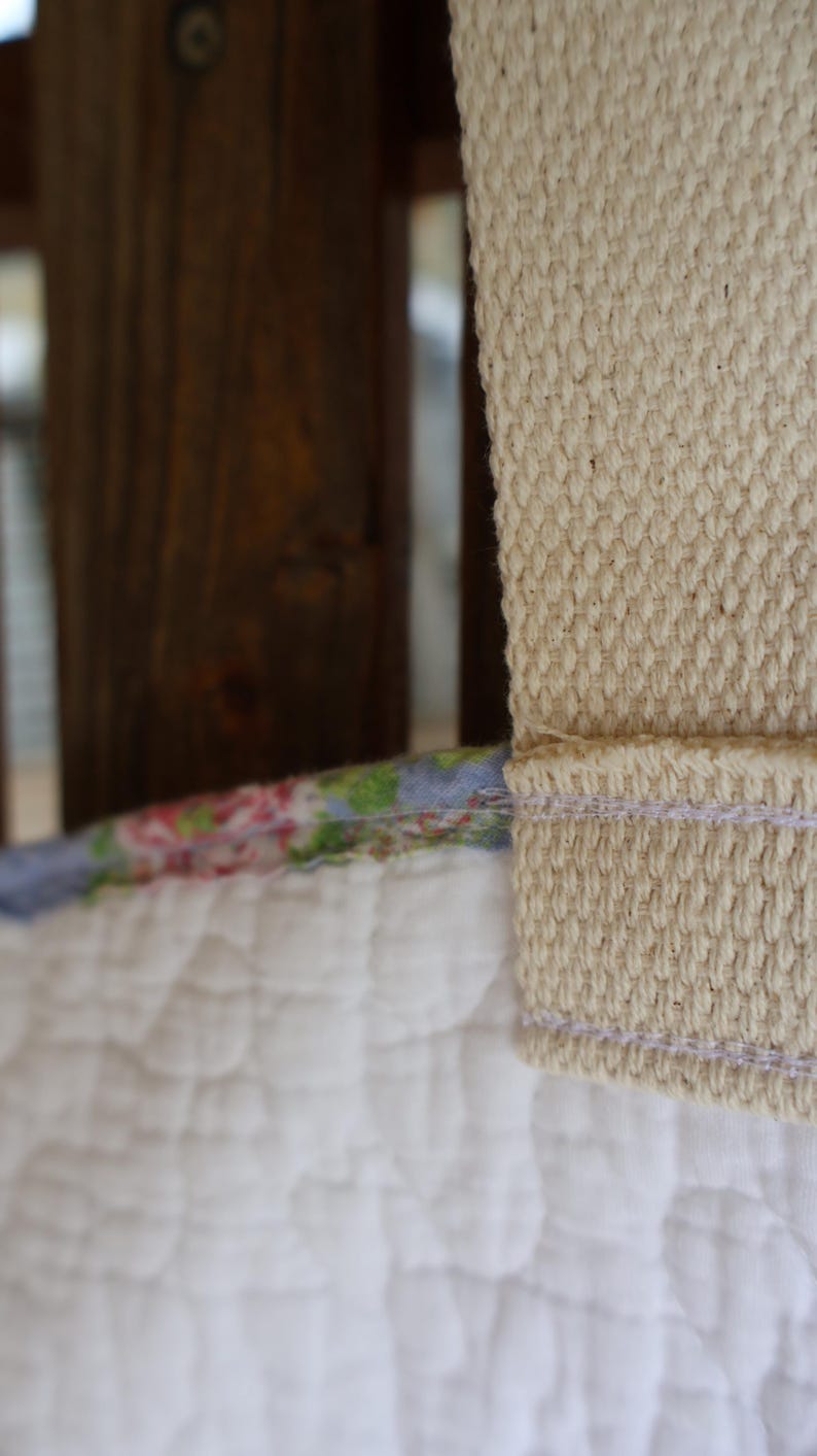 May include: Close-up of a quilted white fabric with a floral patterned edge and a beige woven strap. The strap is stitched to the fabric with a light purple thread. The background features a wooden structure.