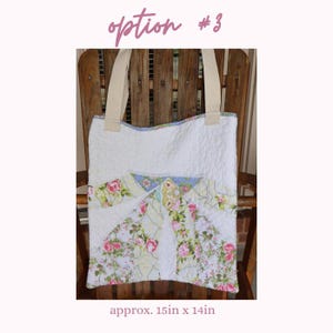 May include: White quilted tote bag with floral patchwork design. Features cream-colored handles and a floral pattern with pink roses and green leaves. The bag measures approximately 15 inches by 14 inches.