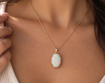 May include: A gold necklace featuring an oval-shaped opal pendant with iridescent flashes of color. The pendant is secured in a simple gold prong setting and hangs from a delicate gold chain, resting against a white ribbed fabric garment.
