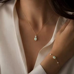 May include: A gold necklace featuring a small round stone above an oval opal pendant, paired with a matching gold bracelet that includes a central oval opal flanked by two small round stones, both set on delicate gold chains against a white shirt.