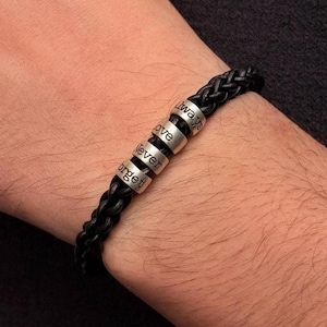 May include: A black braided leather bracelet featuring four cylindrical silver-toned metal beads. Each bead is engraved with a word, collectively forming the phrase Always Love Never Forget, arranged in a vertical sequence along the leather cord.