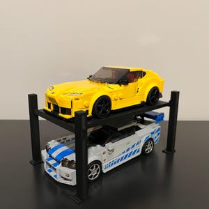 May include: A black metal car lift with two yellow and white toy cars on it. The top car is yellow and the bottom car is white with blue stripes.