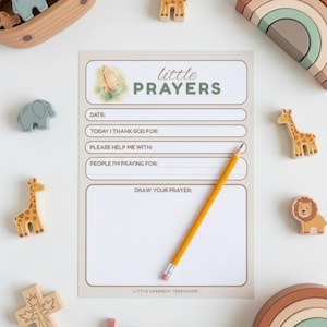 May include: A beige Little Prayers notepad with a pencil, surrounded by wooden animal toys. The notepad features prompts for prayer, including "Today I thank God for" and "Draw your prayer." The notepad has the text "Little Prayers" at the top.