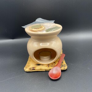 May include: A beige ceramic oil burner with a small candle holder and a wooden spoon. The burner is sitting on a brown fabric mat with a pattern of symbols.
