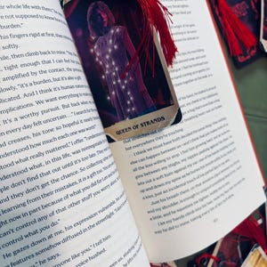 May include: A bookmark with a red tassel and a card featuring an illustration of a person with glowing lights. The text "QUEEN OF STRANDS" is visible on the card. The bookmark is placed on an open book.