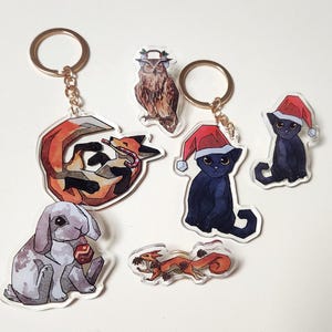 Könnte beinhalten: Eine Sammlung von Schlüsselanhängern mit Tiermotiven. Die Schlüsselanhänger zeigen einen Fuchs, eine Eule, eine Katze, ein Kaninchen und ein Eichhörnchen. Die Katze und die Eule tragen festliche Hüte. Die Schlüsselanhänger haben goldfarbene Ringe.