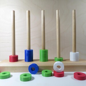 Könnte beinhalten: Ein Holzspielzeug mit fünf Stiften und bunten Ringen. Die Ringe sind rot, grün, blau und weiß. Das Spielzeug steht auf einem Holzsockel.