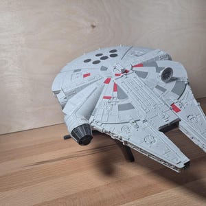 Pode incluir: Um modelo de plástico cinzento de uma nave espacial circular com um casco plano em forma de disco, detalhes mecânicos intrincados na superfície, detalhes retangulares vermelhos, uma antena de radar circular e um motor cinzento escuro num suporte preto.