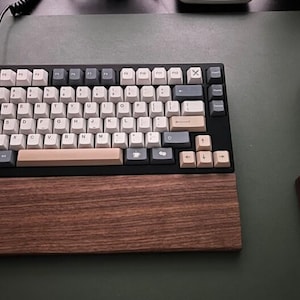 May include: A black and white keyboard with a wooden wrist rest. The keyboard has a light brown keycap set with a few gray and black keys. The keyboard is on a green surface.