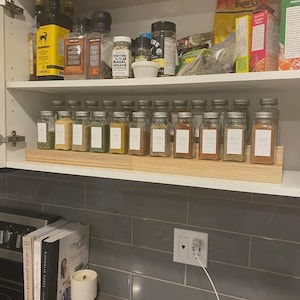 Puede incluir: Un armario de cocina abierto con una especiero de madera que contiene varios frascos de especias de vidrio con etiquetas blancas. El estante superior contiene recipientes de especias adicionales. Se ve un salpicadero de azulejos gris.