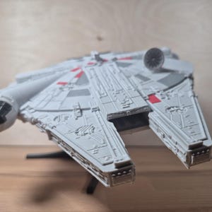 May include: A detailed model of the Millennium Falcon starship, mainly grey with red highlights. The model has intricate paneling and a circular radar dish. The ship is displayed on a black stand, against a light brown background.