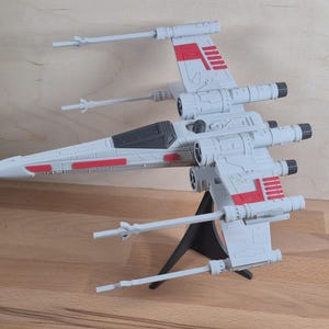 Puede incluir: Un modelo de nave espacial X-wing de plástico gris con marcas rojas en las alas y el fuselaje. La nave cuenta con cuatro cañones láser, una cabina de color gris oscuro y está montada sobre un soporte triangular de color negro.
