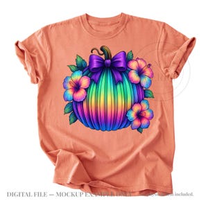May include: A peach-colored t-shirt featuring a central graphic of a pumpkin with a rainbow gradient, topped with a large purple bow and surrounded by colorful hibiscus flowers with green leaves. The design is vibrant and multi-colored.