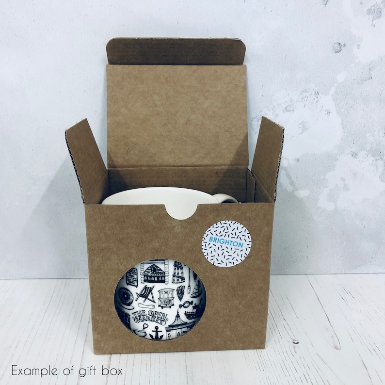 May include: A brown cardboard gift box with a white mug inside. The mug has a black and white design with the word "Brighton" on a blue circle sticker.