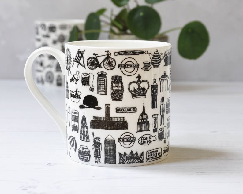 May include: White ceramic mug featuring a black and white illustration of London landmarks, including Big Ben, the London Eye, and a double-decker bus. The mug also features the text "Fish & Chips" and "Oxford Street".