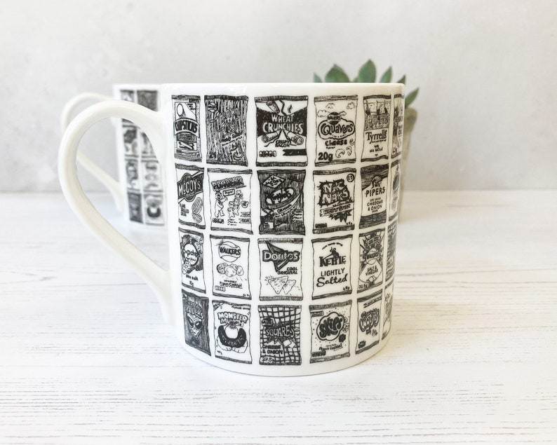 May include: A white ceramic mug with a black and white design of various snack food packaging. The mug has a handle and is printed with the names of popular British snack brands, including McCoy's, Walkers, and Skips.